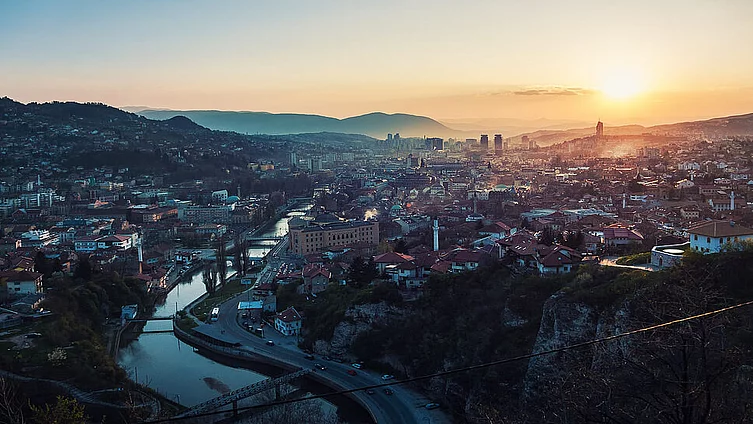 Slika prikazuje zalazak sunca nad Sarajevom, okruženim planinama, s rijekom Miljackom koja teče kroz centar.