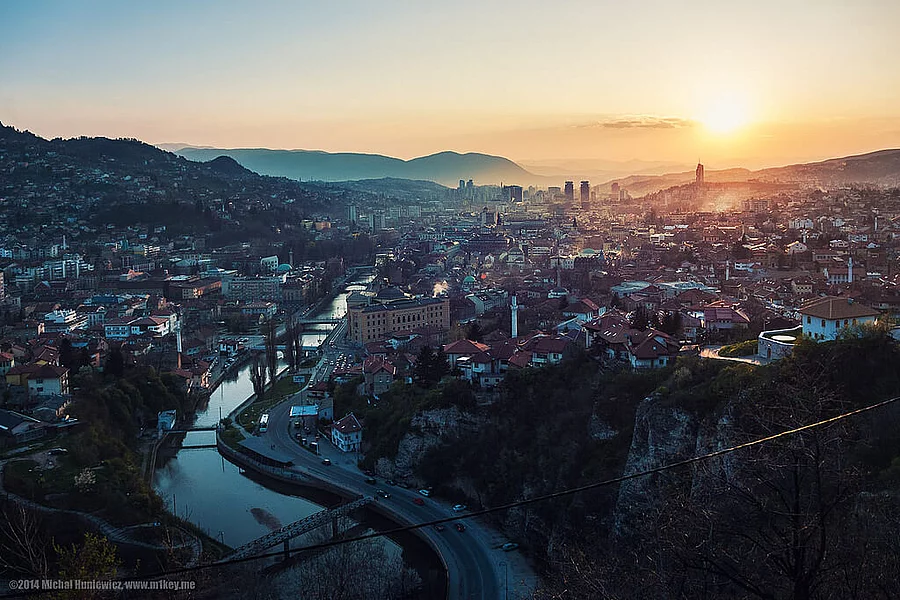Die Aufnahme zeigt einen Sonnenuntergang über Sarajevo, umgeben von Bergen, mit dem Fluss Miljacka, der durch das Zentrum fließt.